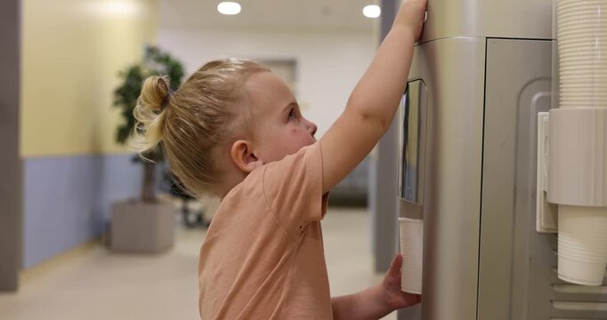 Young independent girl with a ponytail takes a disposable cup and pours fresh, clean water for herself from a cooler, getting her t-shirt wet in the process and quenching her thirst