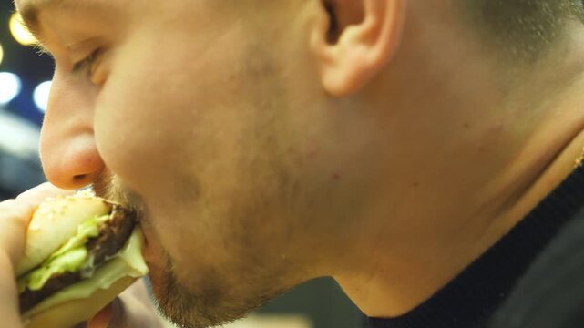 Close up of young man eating hamburger or cheeseburger indoor at evening. Side view of male mouth biting and chewing burger. Slow motion