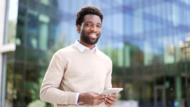 Portrait of smiling african american businessman holding digital tablet standing on street near office building. Happy entrepreneur in casual sweater looking at camera Confident manager posing outside