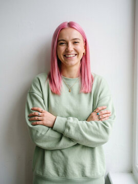 Young woman with pink hair smiling confidently while wearing a light green sweatshirt with her arms crossed indoors portrait