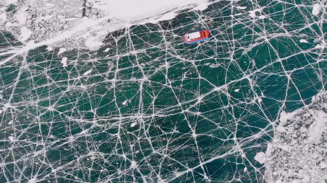 Aerial Of Fractured Emerald Ice Revealing Teal Depths at winter ice lake Baikal with red hovercraft