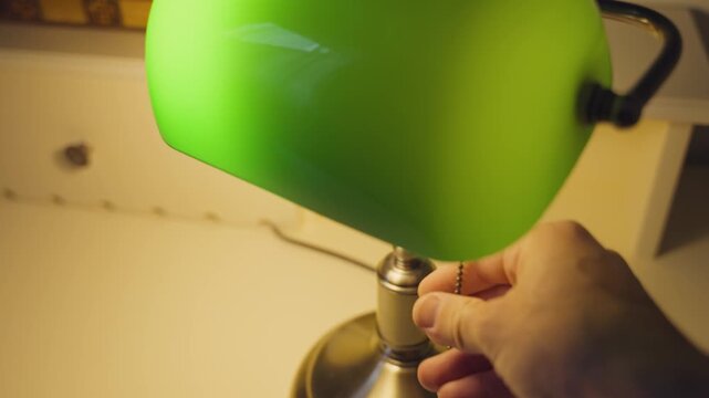 Slow motion footage showing a hand reaching out in a dark room and pulling a chain to turn on a vintage green desk lamp, illuminating the space with a warm and inviting light