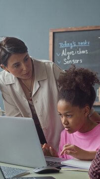 Vertical video of female teacher guiding African American schoolgirl typing on laptop in classroom. Footage is perfect for marketing campaigns focusing on STEM learning and modern education