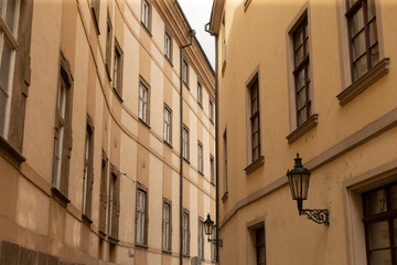A narrow urban alley framed by tall historic beige buildings with multiple windows and decorative stone sills. Ornate black wrought iron wall lamps are © Alena
