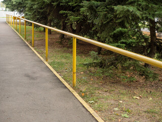 Obraz premium long, yellow metal handrail bordering a paved asphalt walkway, set against a backdrop of mature evergreen trees in a park or public facility