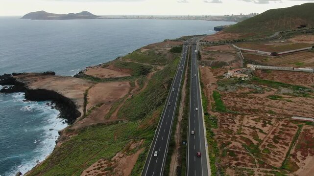 High aerial perspective of cars driving on the Northern Highway along the rugged coastline of Gran Canaria, with the Atlantic Ocean waves crashing