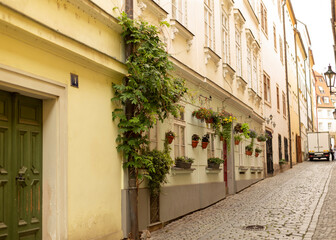 Obraz premium A quiet narrow European street paved with cobblestones lined by historic multi-story buildings. Pale yellow and cream facades feature window boxes and hanging