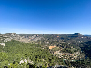 Obraz premium A wide panoramic shot overlooking a majestic canyon covered in lush green pine forests, featuring a winding river and a small rural field under a vast, clear blue sky.