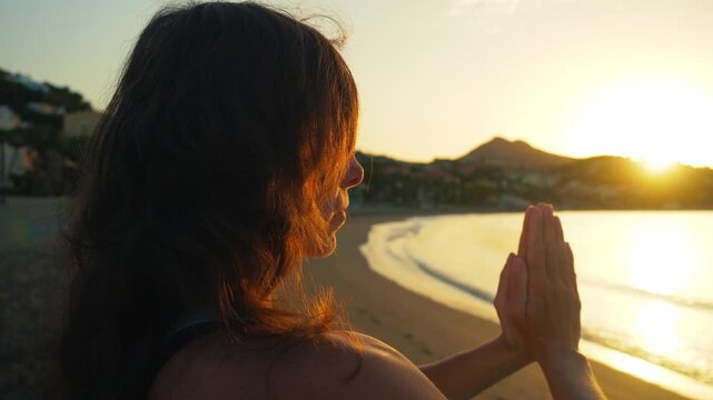 Woman finding inner peace with hands in prayer pose on a serene beach at golden hour sunset, perfect for wellness and travel content.