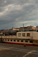 Obraz premium A moored riverboat with a white and red hull sits alongside a stone quay in front of a row of historic multi-story buildings. Festive bunting stretches above