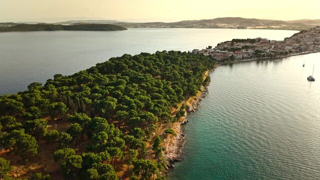 Aerial view of Ermioni peninsula, Greece at sunset. Dense green pine forest bordering a tranquil coastal town with turquoise Aegean Sea waters