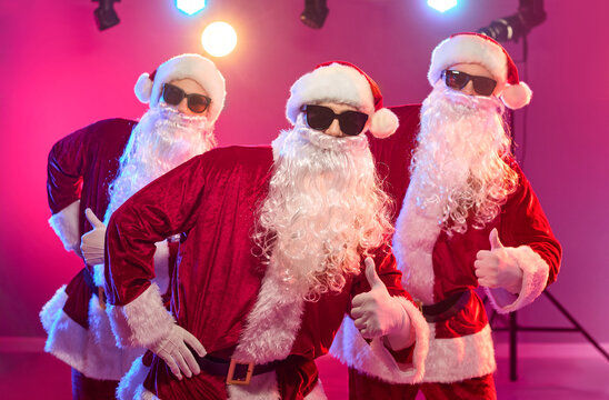 Santas dance at Christmas party in costumes. A trio of Santa dancers gives thumbs up in sunglasses and beards under neon stage lights. Festive holiday party celebration and dance concept.