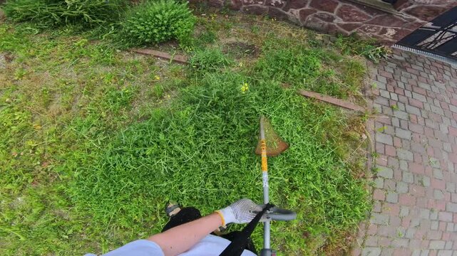 Operator trimming overgrown lawn edge with grass trimmer from overhead operator viewpoint showing tool shaft diagonal toward vegetation and hardscape boundary