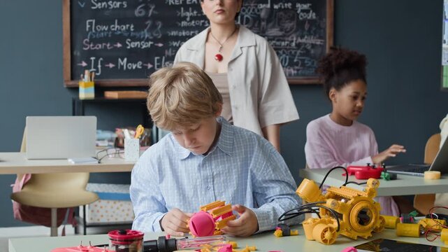 Caucasian kid assembling robot toy during STEM class while female teacher approaches to guide him. Ideal for marketing campaigns promoting STEM education and technology learning programs