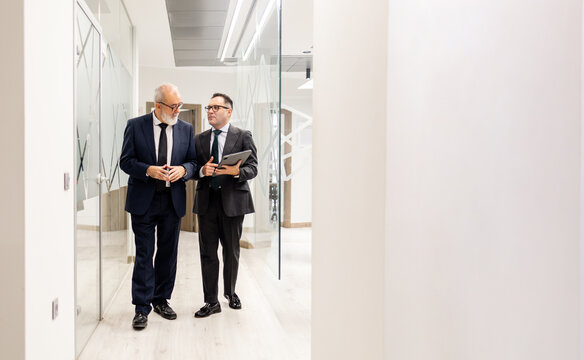 Full shot of two businessmen or lawyers in suits, aged between 50 and 60, walking and talking while looking at a tablet. Concept of executive staff in a modern office with copyspace on the right.
