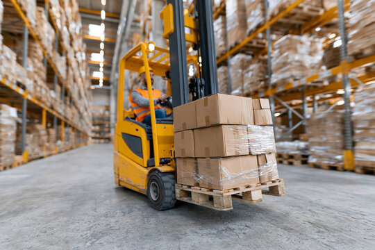 Worker operates forklift in bustling warehouse. Stacked boxes move through industrial space. Yellow machine glides past tall shelves. Bright overhead lights illuminate busy scene