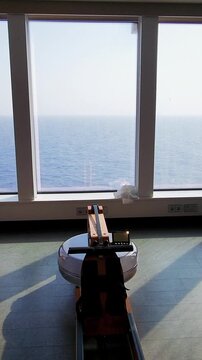 Contemporary fitness room with premium wooden water rowing machines positioned beside panoramic windows overlooking the sea. Bright natural sunlight, modern wellness interior and healthy lifestyle