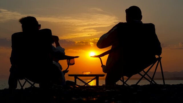 Silhouetted friends enjoying sunset on beach with coffee. Silhouetted friends enjoying tranquil seaside moment, sitting in chairs while sipping coffee against vibrant evening sunset