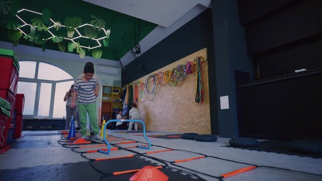 Kids engage in agility training, utilizing cones and hurdles for fitness exercises. Youngsters develop physical coordination and motor skills in a playful indoor setting.