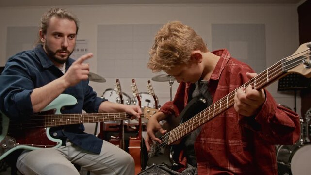 Young male music teacher instructing teenager on playing bass guitar in studio. Ideal for music school marketing and private tutoring ads