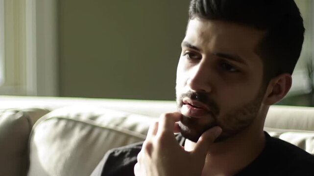 Thoughtful man sitting on sofa, resting chin on hand, pensive expression, soft natural light.