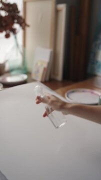 Slow motion closeup of a painter's hand using a spray bottle to wet a blank canvas, preparing the surface for watercolor or acrylic painting in an art studio, showing creative process. vertical