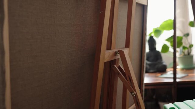 Close-up of a blank canvas resting on a wooden easel inside a naturally lit art studio, with a blurred background showing a window, a plant, and a small Buddha statue on a shelf