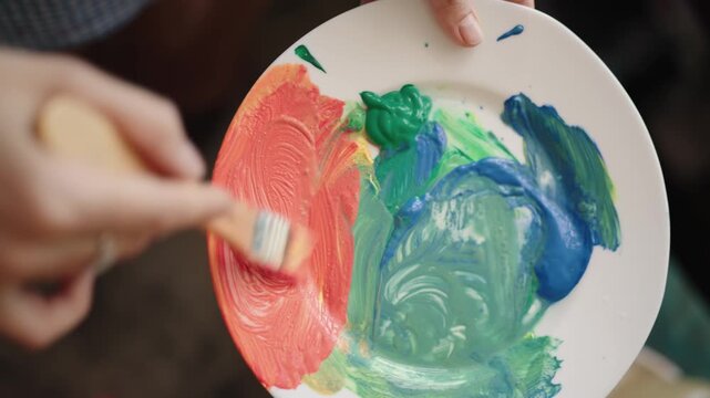 Close-up shot of an artist's hand holding a ceramic plate used as a palette, mixing vibrant red, green, and blue acrylic paints with a paintbrush in slow motion for a new creation