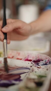 Close-up of an artist's hand holding a brush and mixing colorful watercolor paints on a white palette in slow motion, showing the creative process in an art studio or workshop. vertical