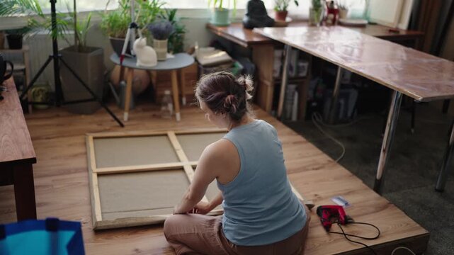 Creative woman artist sitting on the floor of her bright studio, carefully stretching and stapling a new large canvas to a wooden frame in preparation for her next masterpiece.