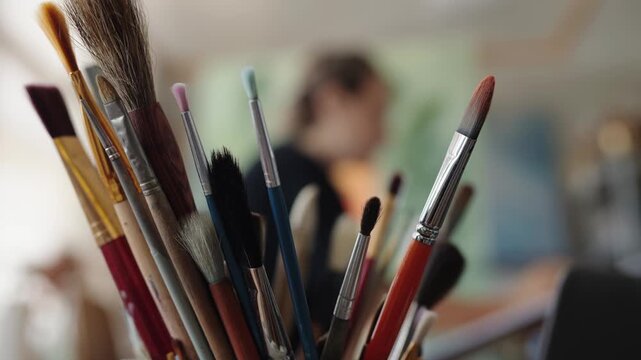Close up of a jar full of paintbrushes in an artist's studio, with the painter working on a canvas in the blurry background. Creative process in a workshop with professional art supplies