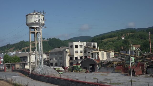 Wide view of an industrial factory complex with water tower, processing buildings, conveyors, and production facilities. Manufacturing site surrounded by green hills in northern Spain.
