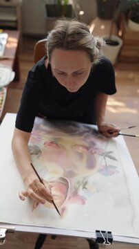 Slow motion, close up view of a woman's hand painting a watercolor on paper with a brush, creating an abstract illustration in a creative workshop with natural light from a window. vertical