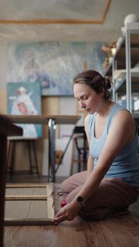 Creative woman artist sitting on the floor of her bright studio, Slow-motion close-up of a female artist stretching and stapling a canvas onto a wooden stretcher bar in her workshop. vertical