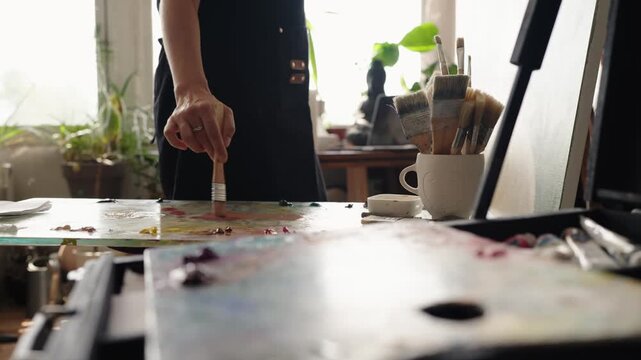 Slow motion of a female painter in a black apron mixing oil paints with a brush on a glass palette, preparing colors for a new masterpiece in a sunlit art studio workshop