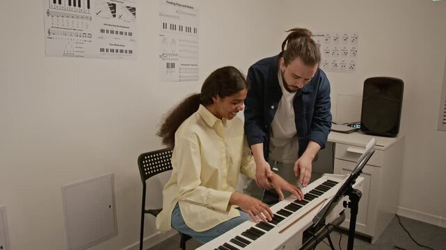 Caucasian young man teaching Black teenager how to play digital piano in music classroom. Footage for promoting music schools, private tutoring, and creative education