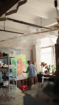 Long shot of a professional female painter creating a modern artwork on a large canvas, using a paintbrush and paints in her sunlit workshop filled with art supplies and plants. vertical