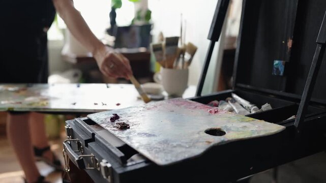 Close-up shot of a female painter's hand using a paintbrush to mix colorful oil paints on a wooden palette in a bright art studio, with an easel and art supplies in the background