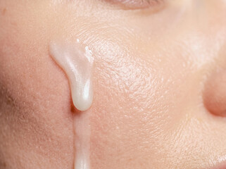 Fototapeta premium Macro close up of a white cosmetic cream lotion drop dripping down a smooth clean woman cheek skin surface. Skincare product application and moisturizing beauty concept.