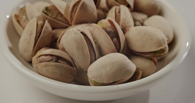 Close-up of roasted and salted pistachios in their shells slowly rotating in a white bowl, a delicious and healthy snack food rich in protein and nutrients, isolated on white background