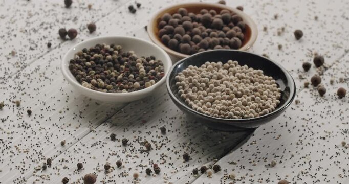 Slow motion footage showing different types of peppercorns falling into small ceramic bowls on a white wooden table, including allspice, and white, black, green and red pepper blend