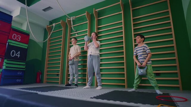 Three children perform kettle bell exercises in a bright gymnasium. These young kids are focused on their physical fitness and strength training.