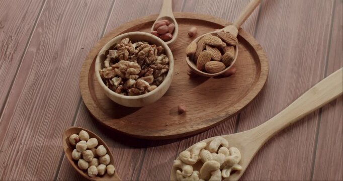 Rotating overhead view of a healthy mix of nuts and seeds, including walnuts, almonds, cashews, hazelnuts, and peanuts, displayed in wooden bowls and spoons on a rustic table background