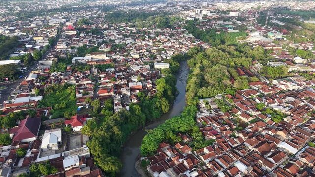 Aerial photo of Jember showing a narrow river winding through dense red-tiled residential neighborhoods, pockets of trees and urban greenery, highlighting riverside living, community layout