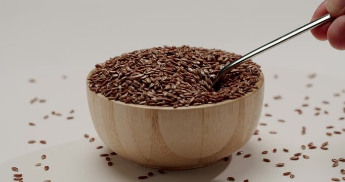 Metal spoon stirring and scooping whole brown flax seeds from a light wooden bowl, with the healthy superfood grains falling in slow motion, all isolated on a clean white background