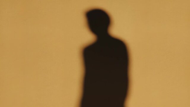 Shadow of a person bending forward against a warm yellow wall, showing gradual movement and posture change in three sequential frames of a video