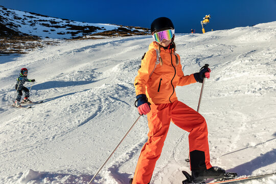 Two siblings ski on bright mountain slope in colorful winter gear, helmets, goggles, and gloves beneath clear blue sky. Family adventure conveys motion, confidence, safety, sport, and alpine fun