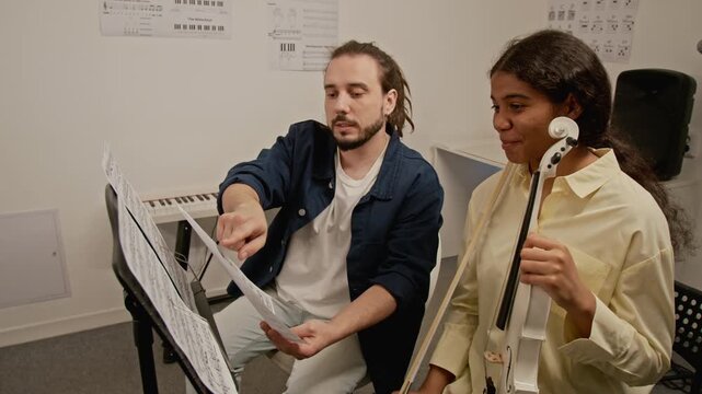 Caucasian male teacher explaining sheet music to African American female student holding violin. Ideal for promoting music schools, private tutoring, and creative education programs