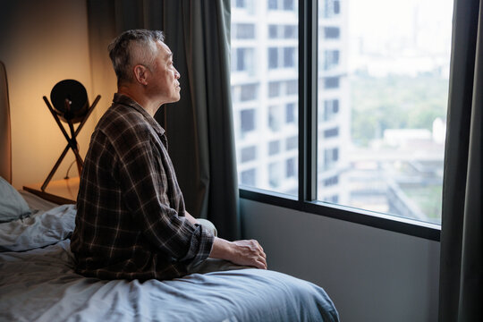 Elderly man contemplates city view from his bedroom window.