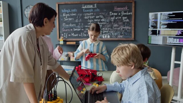 Female teacher guiding Caucasian boy typing on laptop while multiethnic kids assemble robots in classroom. Footage is ideal for promoting STEM education and modern school programs